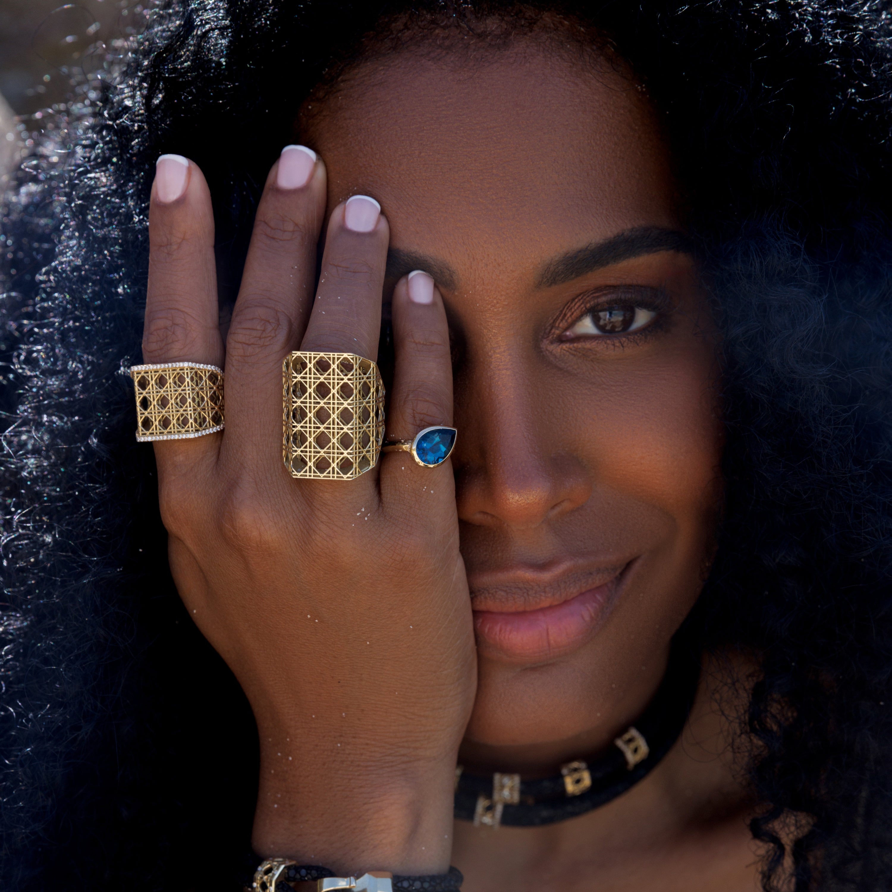 MoAnA LUU Doudou 18K gold rings with woven cane design and Dlo pear-shaped London blue topaz ring worn on model, showcasing sculptural Creole-inspired jewelry.