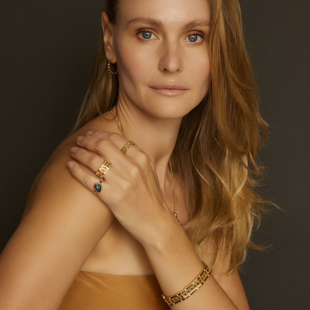 MoAnA LUU fine jewelry worn on model, featuring Doudou gold rings with woven design, Dlo pear-shaped blue sapphire ring, Ti hoops and Doudou gold bracelet in 18K yellow gold inspired by Creole craftsmanship.
